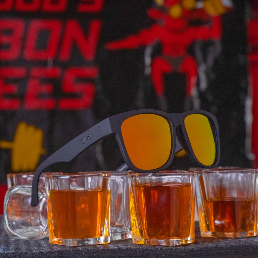Óculos de sol unissex preto com armação quadrada Beelzebub's Bourbon Burpees em foco sob copos.