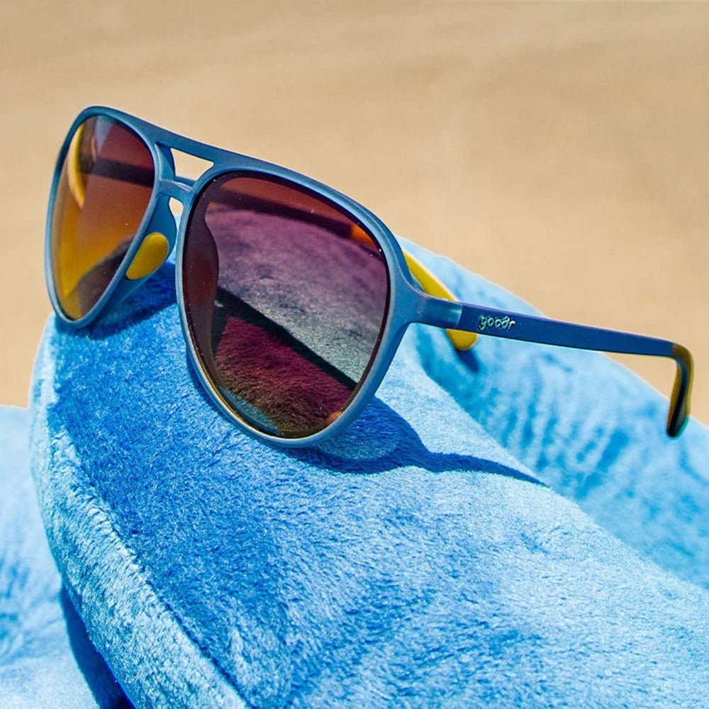 Óculos de sol Goodr Brasil modelo aviador com armação azul e lentes amarelas degradê sobre almofada de viagem azul.