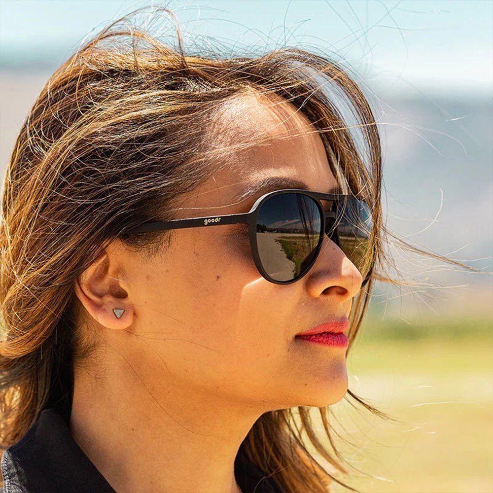 Mulher usando óculos de sol Goodr Brasil modelo aviador preto com lentes polarizadas em ambiente externo ensolarado.