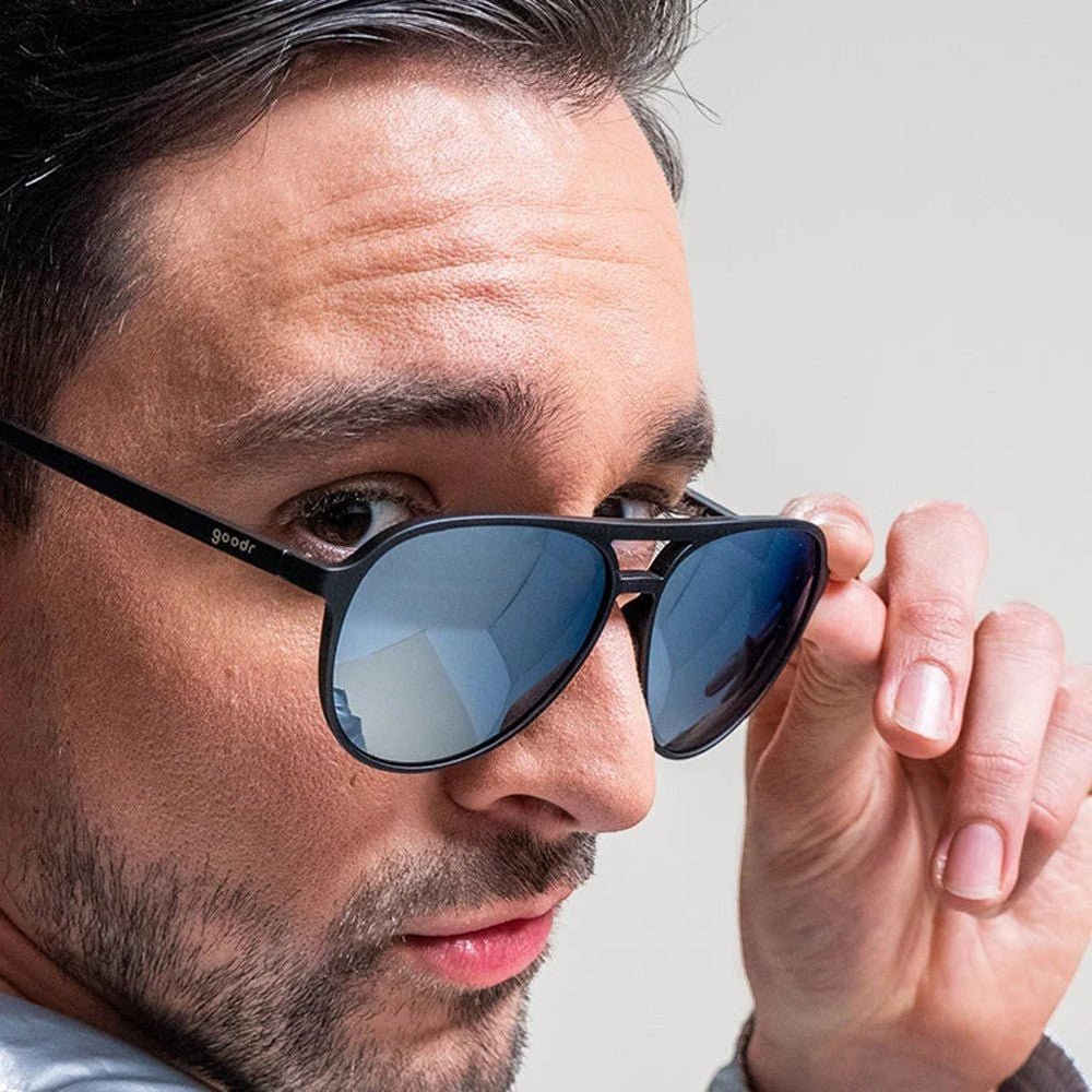 Homem usando óculos de sol aviador Goodr Brasil preto com lentes cinza espelhadas e design moderno.