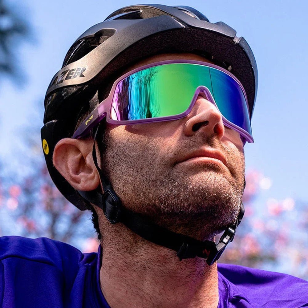 Ciclista usando óculos Goodr Brasil roxos com lente única verde espelhada e capacete, ideal para esportes ao ar livre.