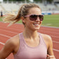 Mulher loira correndo em pista de atletismo usando óculos de sol Goodr Brasil com armação rosa e lentes espelhadas.