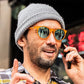 Homem usando óculos de sol Goodr Brasil laranja translúcido com lentes azuis espelhadas e gorro cinza em ambiente urbano