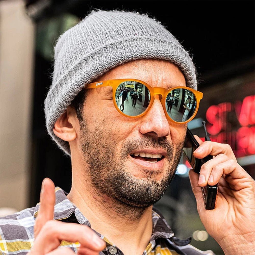 Homem usando óculos de sol Goodr Brasil laranja translúcido com lentes azuis espelhadas e gorro cinza em ambiente urbano