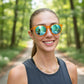 Mulher sorridente usando óculos de sol Goodr Brasil com armação laranja translúcida e lentes azuis espelhadas.