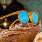 Óculos de sol Goodr Brasil modelo Circle G com armação laranja translúcida e lentes azuis espelhadas sobre um pão.