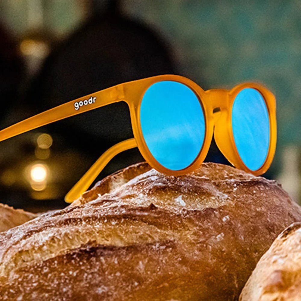 Óculos de sol Goodr Brasil modelo Circle G com armação laranja translúcida e lentes azuis espelhadas sobre um pão.