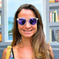 Mulher usando óculos de sol Goodr Brasil redondo com armação transparente e lentes roxas espelhadas.