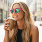 Mulher loira sorrindo usando óculos de sol Goodr Brasil com armação rosa e lentes azuis espelhadas em ambiente urbano