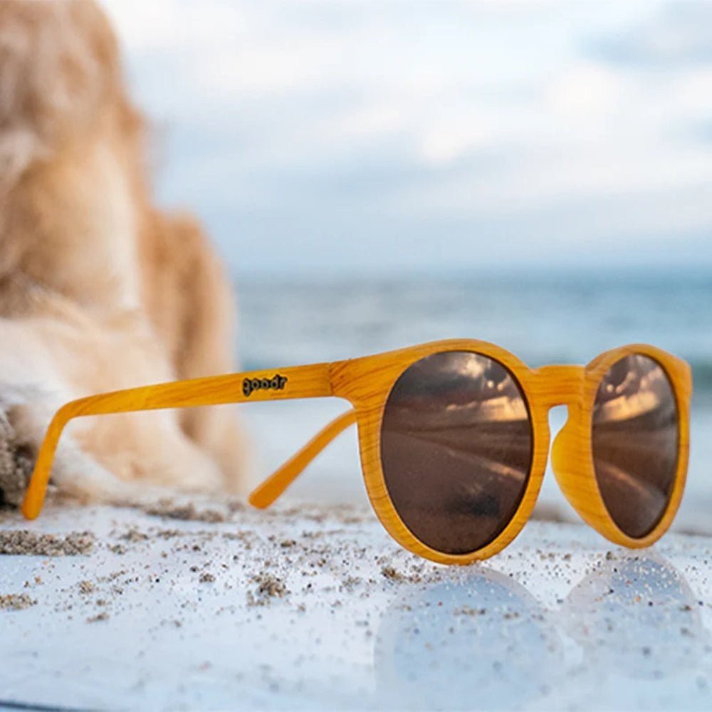 Óculos de sol Goodr Brasil Bodhi's Ultimate Ride com armação caramelo amadeirada e lentes marrons na areia da praia.