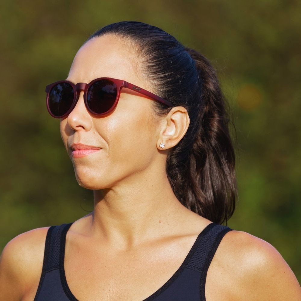 Mulher usando óculos de sol Goodr Brasil modelo Circle G bordô com lentes marrons polarizadas em ambiente externo.