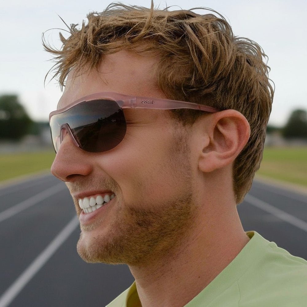 Homem sorrindo usando óculos de sol esportivos Goodr Brasil com armação translúcida e lentes polarizadas em pista.