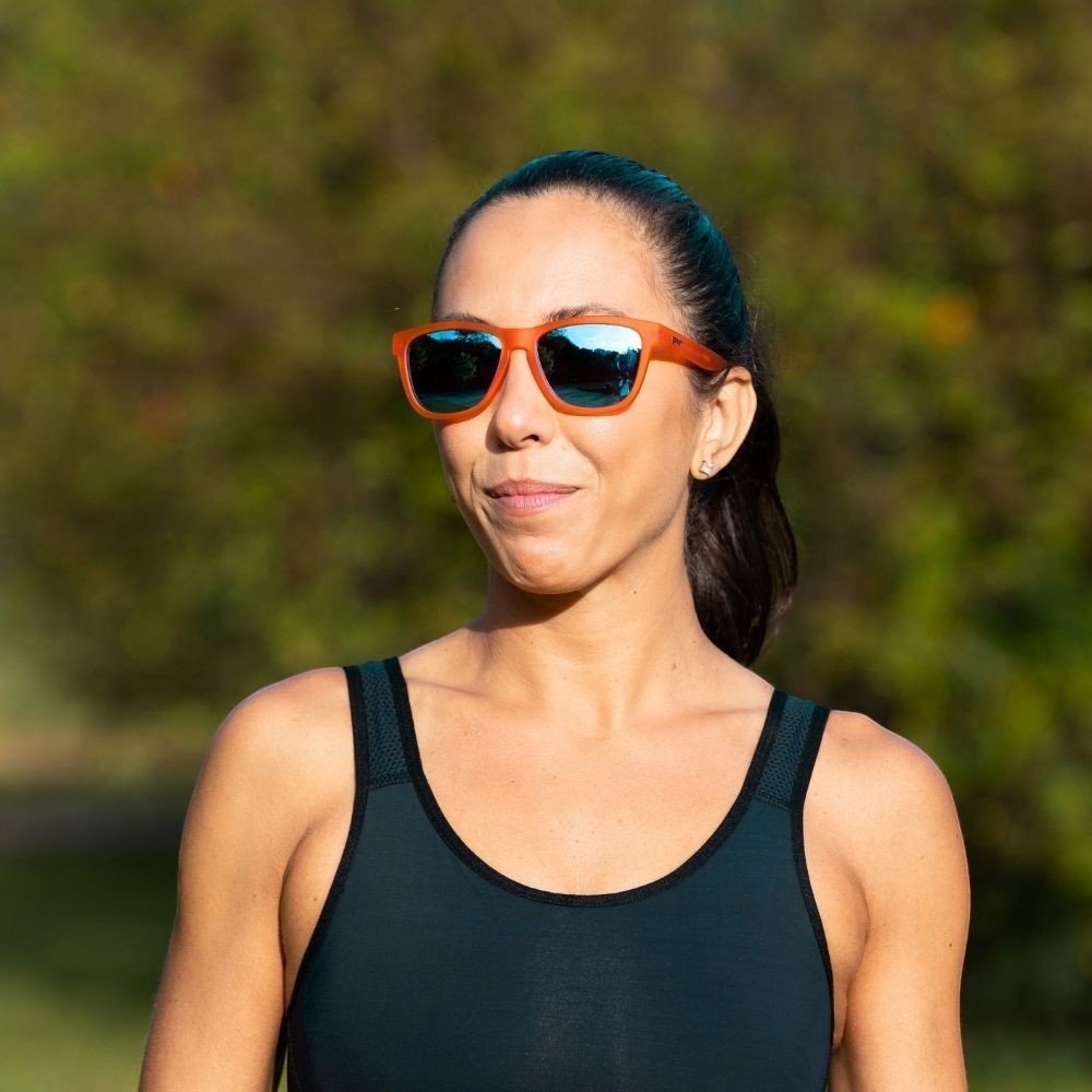 Mulher usando óculos de sol Goodr Brasil Donkey Goggles com armação laranja e lentes azuis espelhadas ao ar livre.