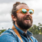 Homem de barba usando óculos de sol Goodr Brasil redondo com armação laranja translúcida e lentes azuis espelhadas.