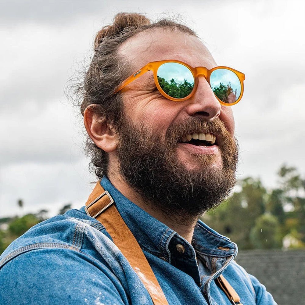 Homem de barba usando óculos de sol Goodr Brasil redondo com armação laranja translúcida e lentes azuis espelhadas.