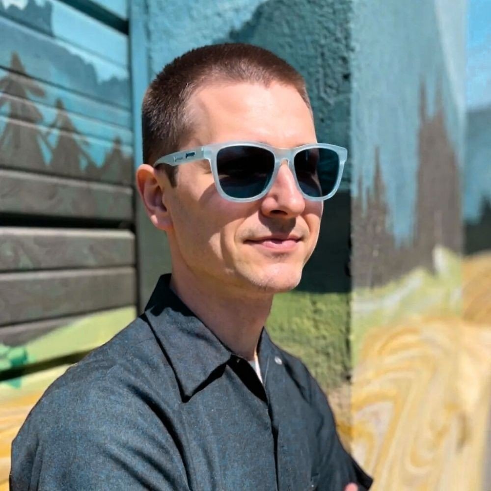 Homem usando óculos de sol Goodr Brasil modelo These Shades Are Still Trash com armação azul claro e lentes escuras.