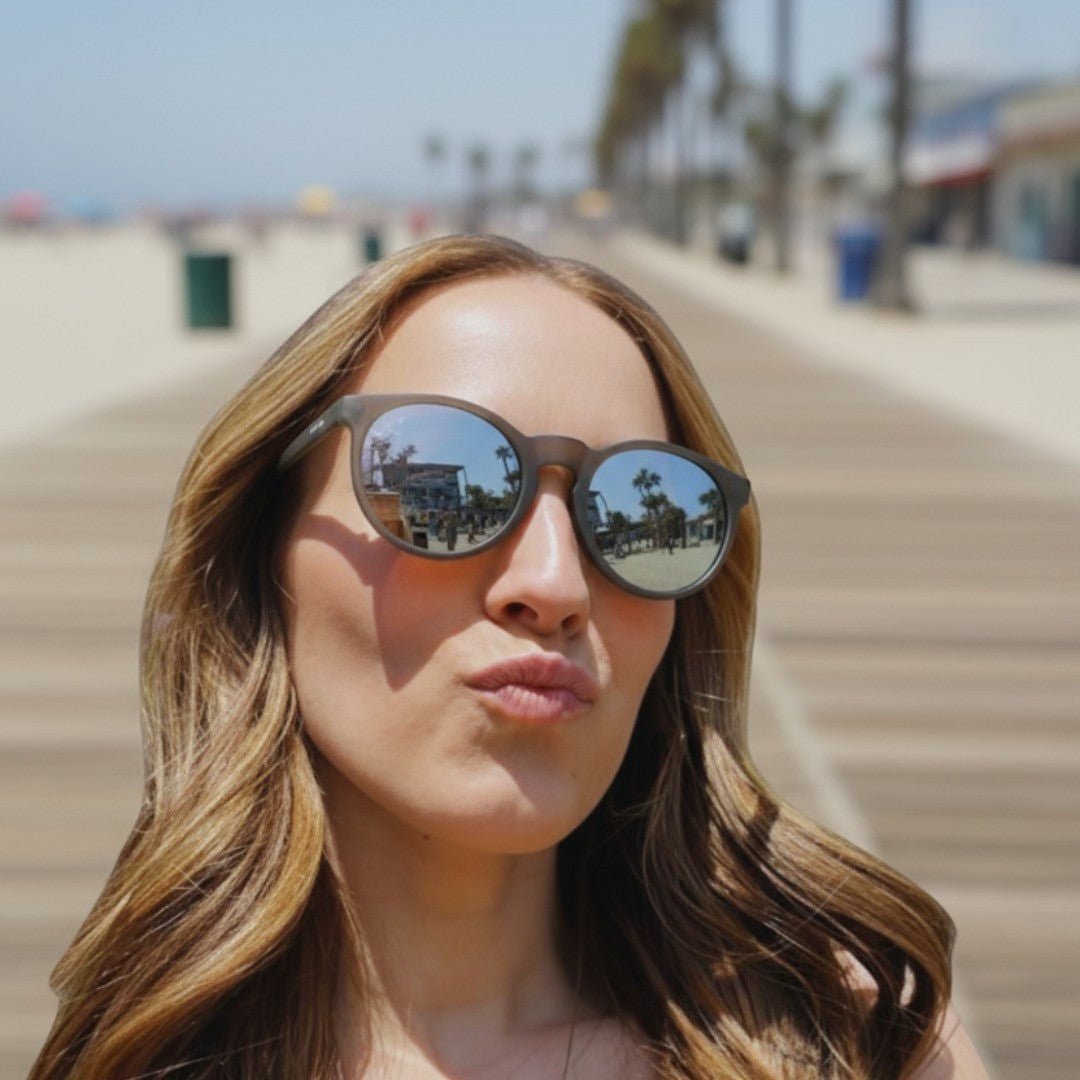 Mulher usando óculos de sol Goodr Brasil They Were Out redondo com armação cinza e lentes espelhadas na praia