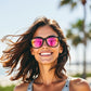 Mulher sorridente usando óculos de sol Goodr Brasil Professional Respawner com armação preta e lentes rosa espelhadas.