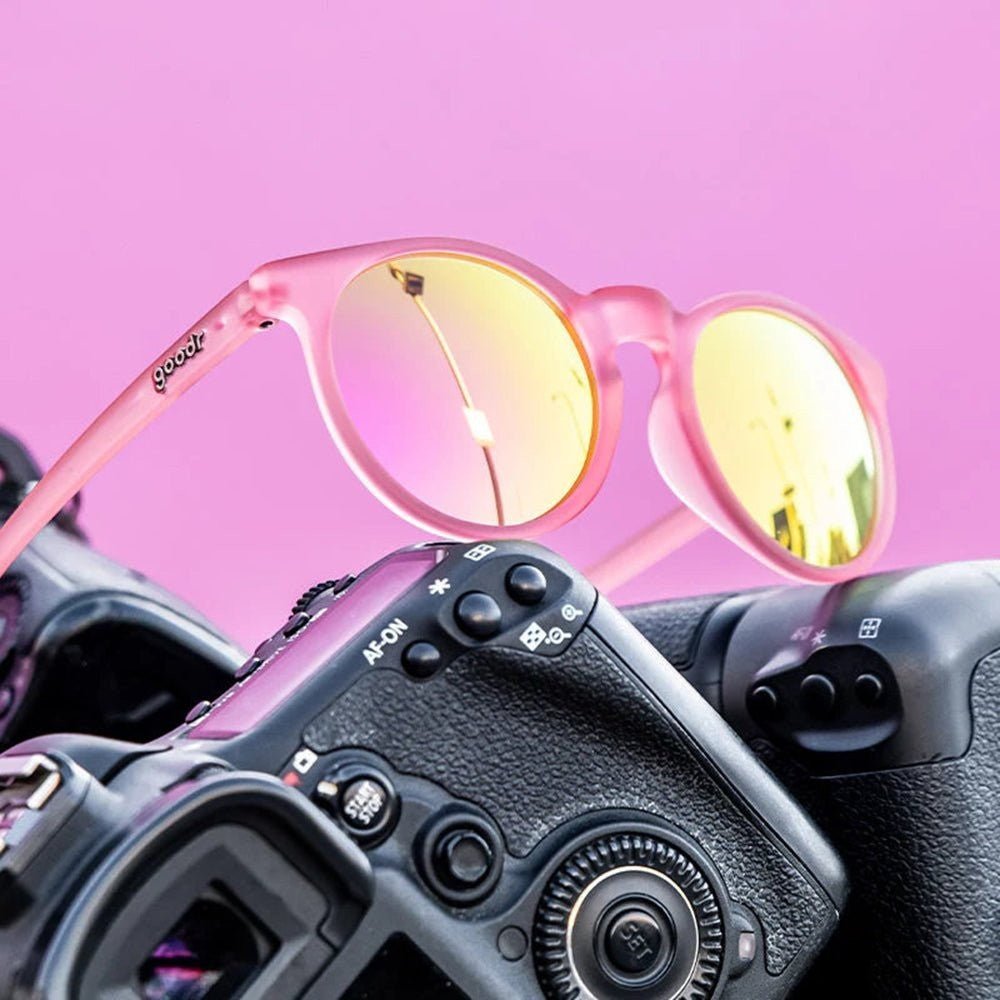 Óculos de sol Goodr Brasil modelo Influencers Pay Double com armação rosa fosca e lentes espelhadas sobre uma câmera