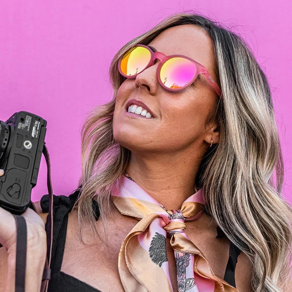 Mulher usando óculos de sol Goodr Brasil Influencers Pay Double rosa com lentes espelhadas em fundo rosa vibrante.