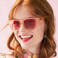 Modelo ruiva sorrindo usando óculos de sol Goodr Brasil modelo Don't Make Me Blush com armação e lentes rosa.