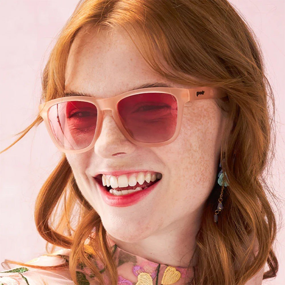 Modelo ruiva sorrindo usando óculos de sol Goodr Brasil modelo Don't Make Me Blush com armação e lentes rosa.
