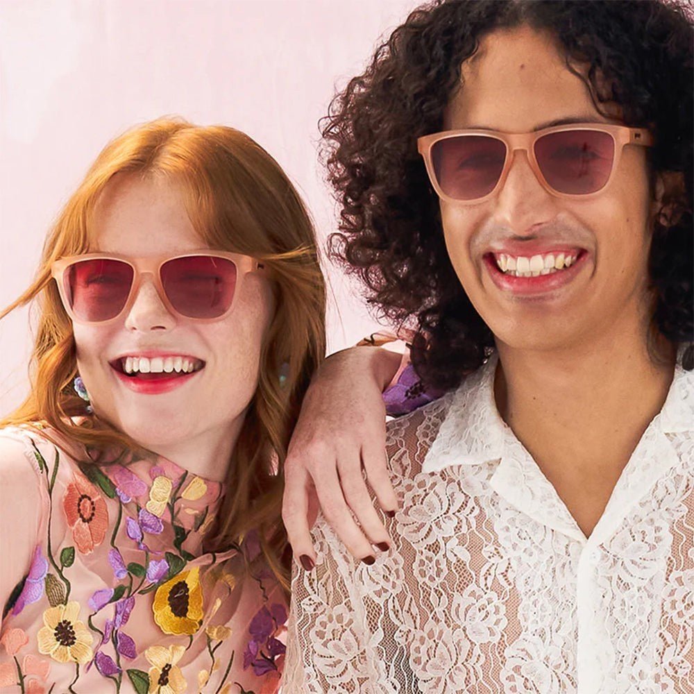Homem e mulher sorridentes usando óculos de sol Goodr Brasil Don't Make Me Blush com armação e lentes rosa.