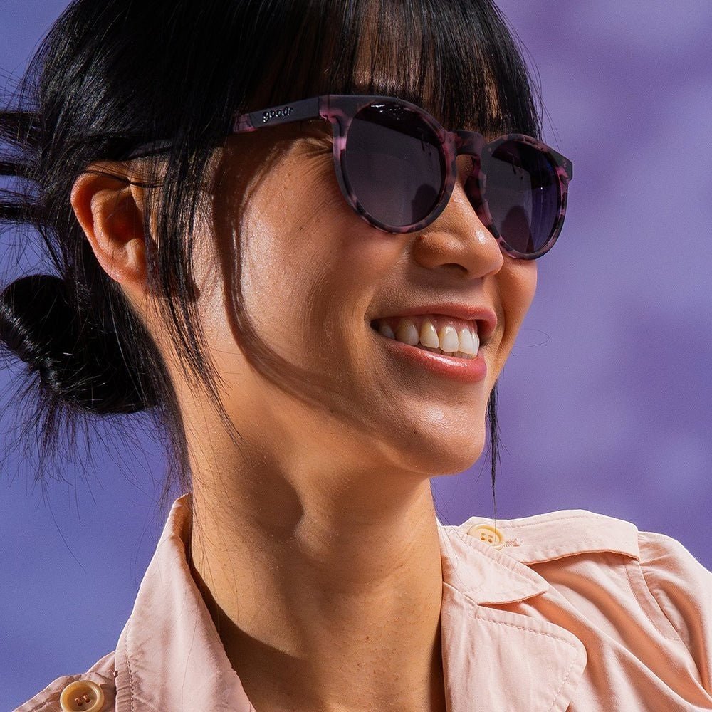 Modelo usando óculos de sol Goodr Brasil Fluent In Silence com armação rosa estampada e lentes pretas polarizadas.