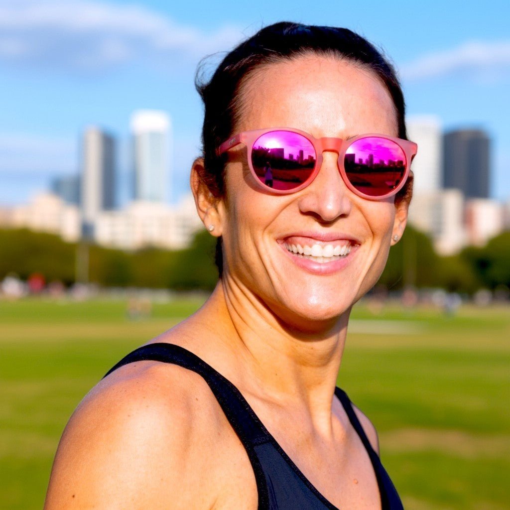 Mulher sorridente usando óculos de sol Goodr Brasil redondo rosa com lentes espelhadas em um parque ao ar livre.