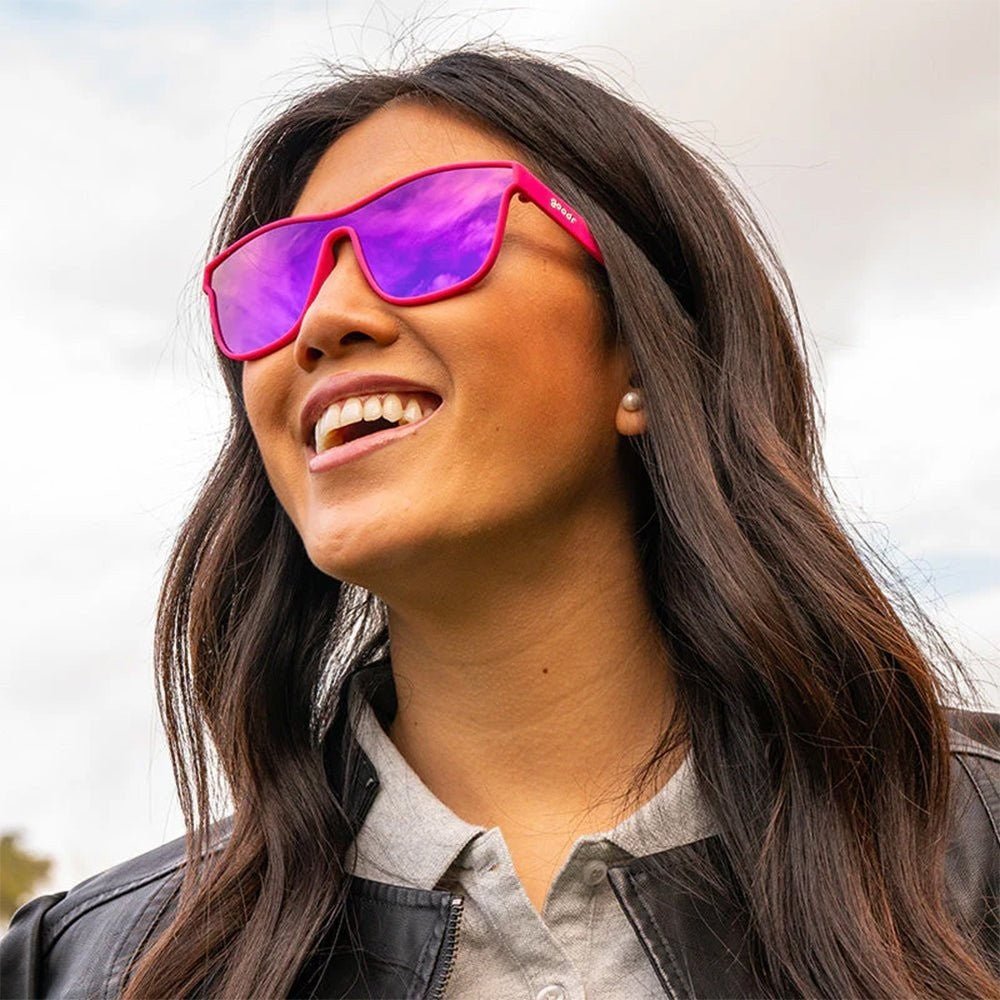 Mulher sorrindo usando óculos de sol Goodr Brasil VRG com armação rosa e lentes roxas espelhadas polarizadas