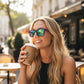 Mulher loira usando óculos de sol Goodr Brasil com armação roxa e lentes verdes espelhadas, tomando café em uma mesa.