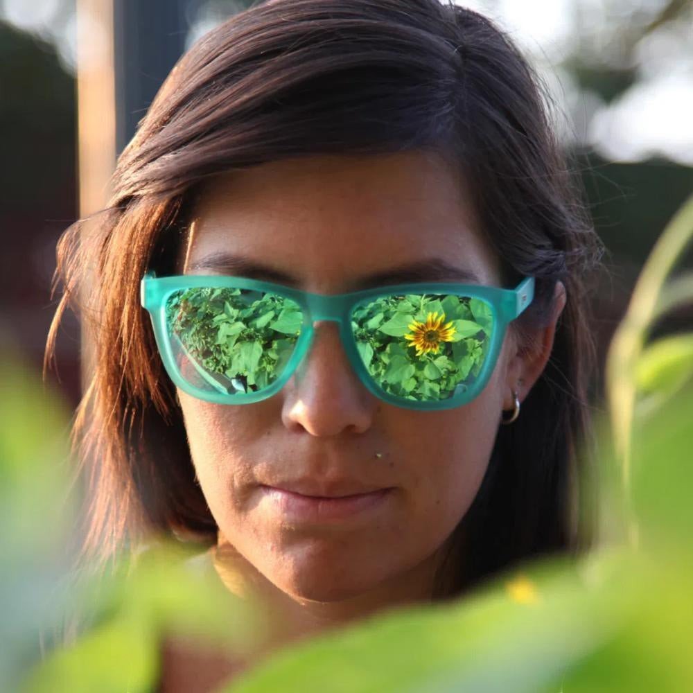 Mulher usando óculos de sol Goodr Brasil verde com lentes espelhadas polarizadas em ambiente externo com girassóis.