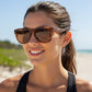 Mulher sorridente usando óculos de sol Goodr Brasil BFG com armação tartaruga e lentes marrons na praia.