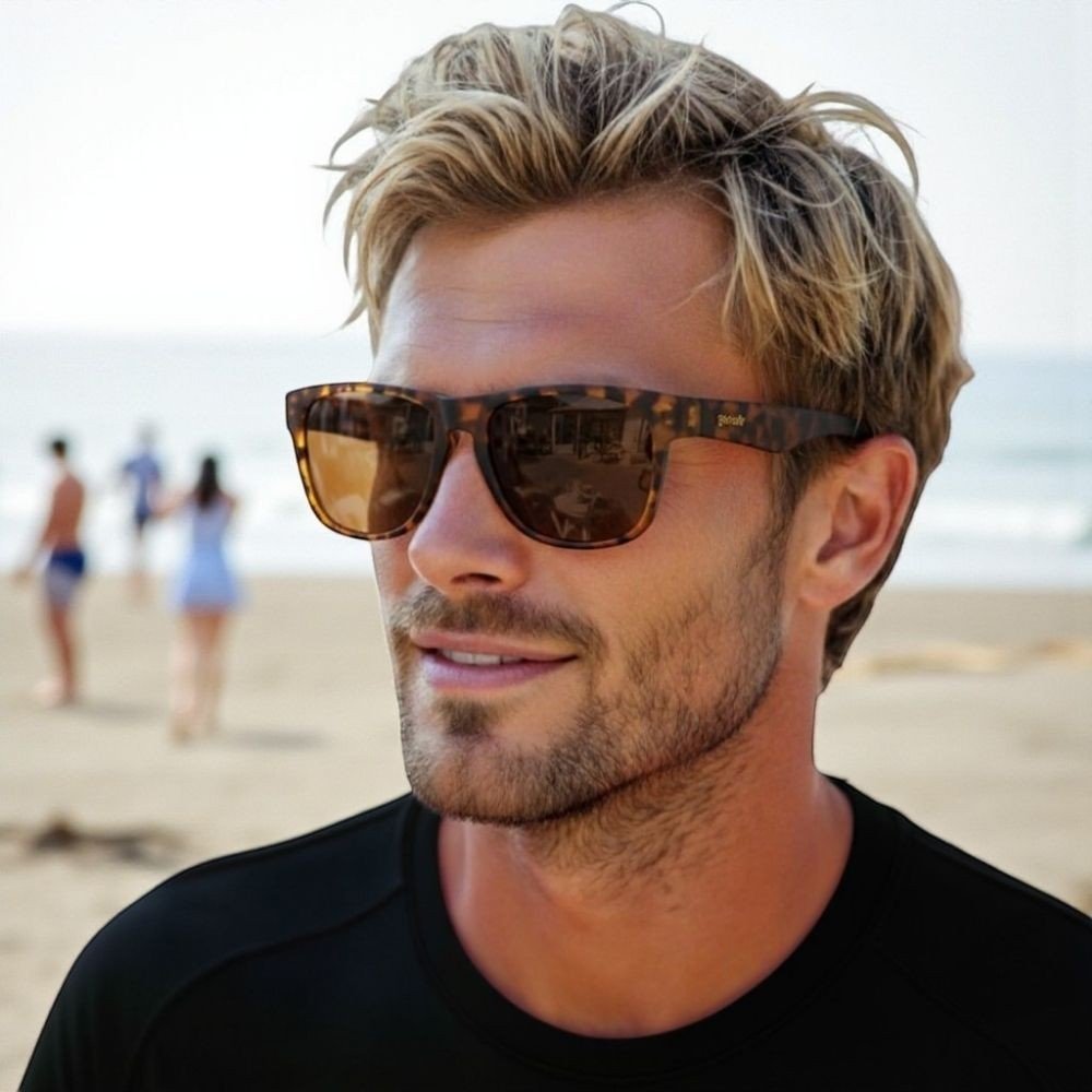 Homem loiro usando óculos de sol Goodr Brasil Hellhound Hallucinations com armação tartaruga e lentes marrons na praia