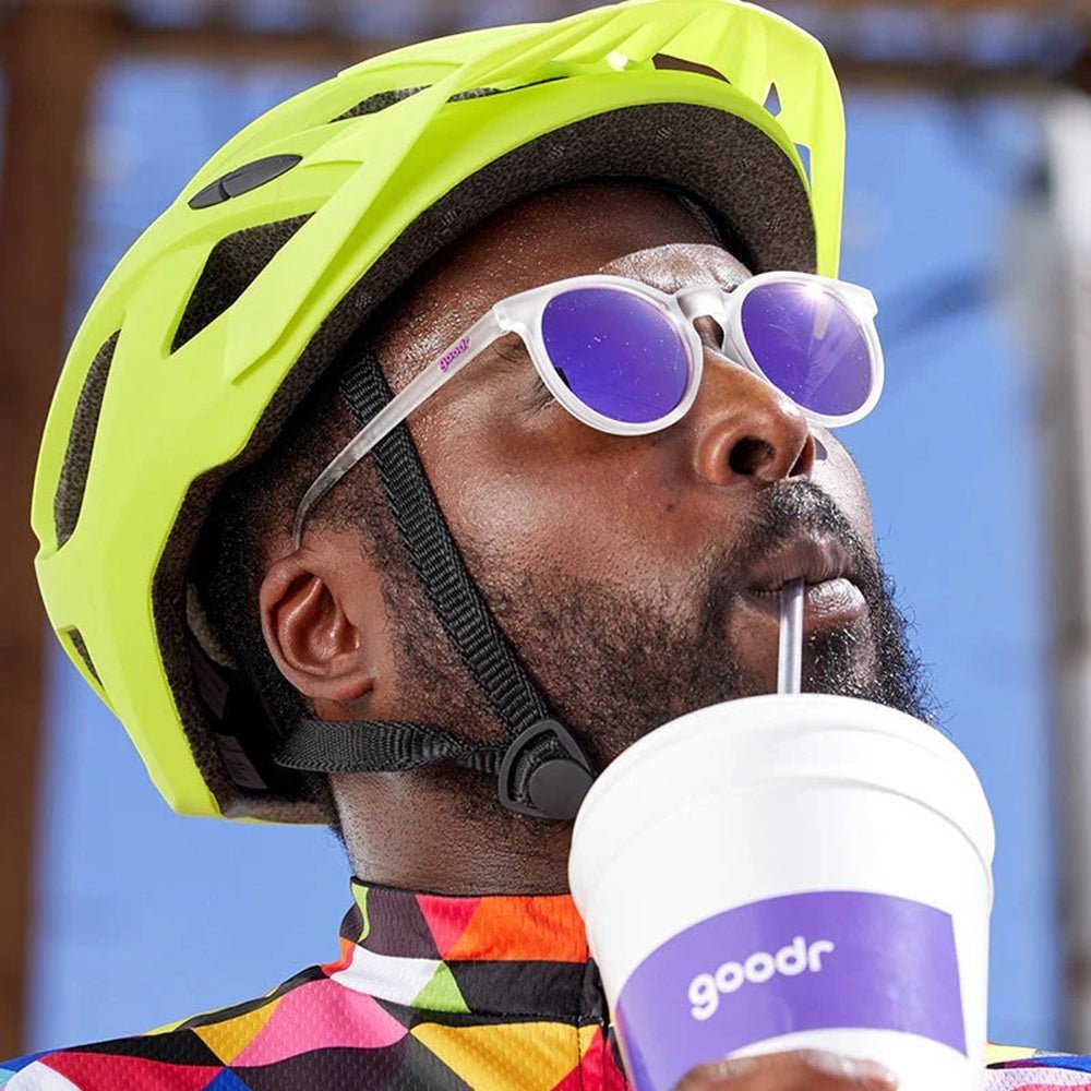 Ciclista usando óculos de sol Goodr Brasil com armação transparente e lentes roxas espelhadas, capacete amarelo e camisa colorida.