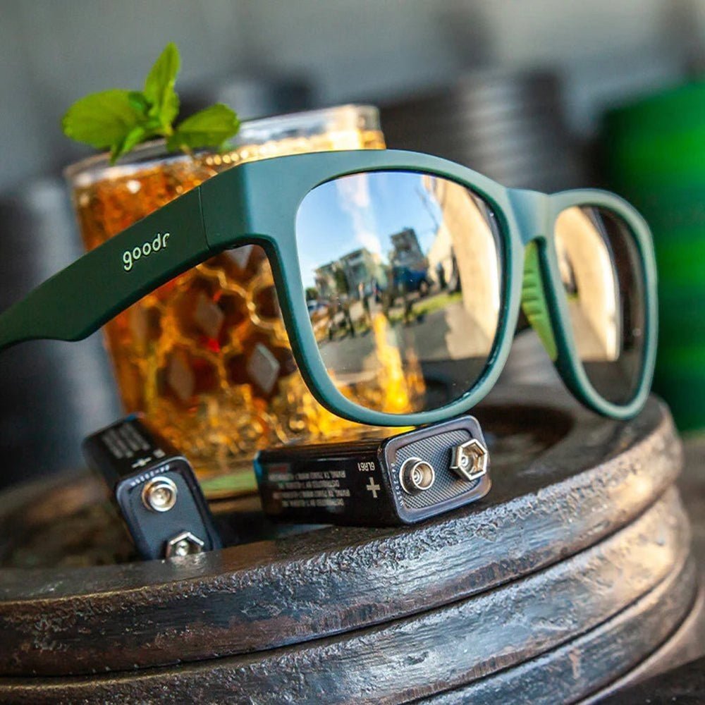 Óculos de sol Goodr Brasil BFG verde com lentes espelhadas polarizadas, apoiado sobre pesos de academia e drink.